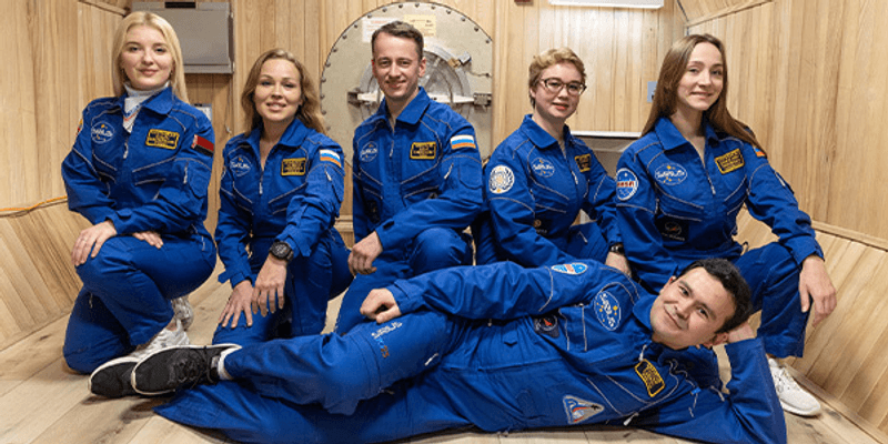 Six people in blue space suits in front of a wooden wall with a hatch behind them, one man laying down in front of the others who are squatting in a "paint me like your french girls" pose