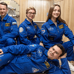 Six people in blue space suits in front of a wooden wall with a hatch behind them, one man laying down in front of the others who are squatting in a "paint me like your french girls" pose