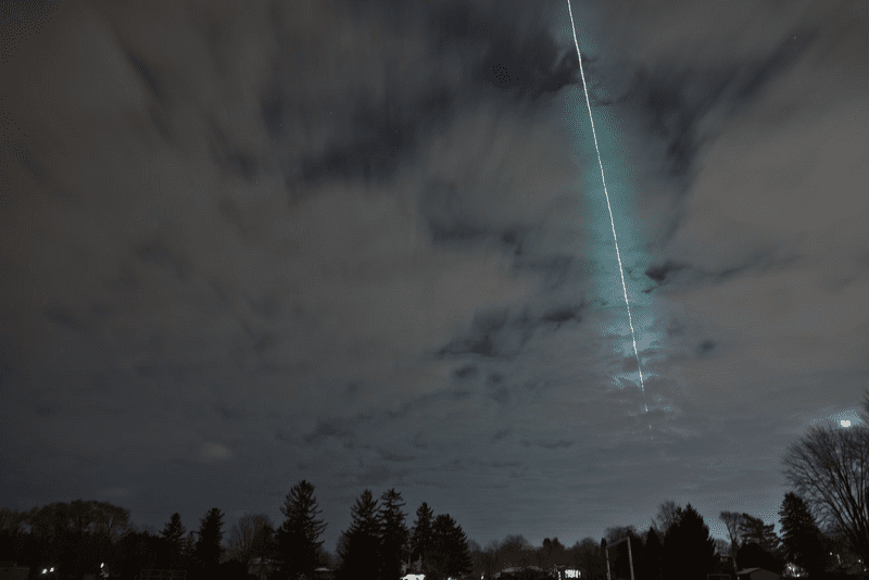 A slightly cloudy sky is photographed. Through it a bright greenish light show the path the the asteroid took as it burned. Trees are visible at the bottom of the frame.