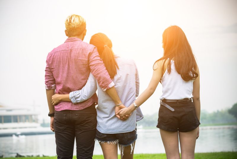 A couple with their arms around each other, but the man is holding hands with another woman behind his partner's back