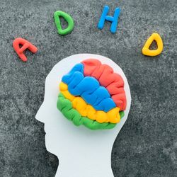 paper cutout of a human head with blue, red, green and yellow plasticine in the shape of a brain; the letters A D H and D are spelled out in plasticine above on a grey stone surface