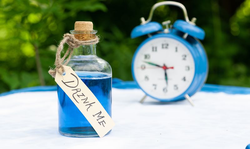 blue liquid in glass bottle labeled "drink me" with blue alarm clock in the background