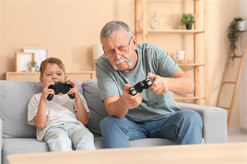 The photo shows an older grandfatherly man playing a Playstation game with is young boy, presumably his grandson. The two are sat on a stone grey sofa in a living room. The man, who is wearing glasses and has a chinless beard, is leaning slightly to his right as he is playing. 