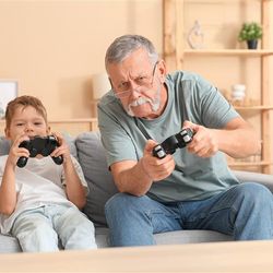 The photo shows an older grandfatherly man playing a Playstation game with is young boy, presumably his grandson. The two are sat on a stone grey sofa in a living room. The man, who is wearing glasses and has a chinless beard, is leaning slightly to his right as he is playing. 