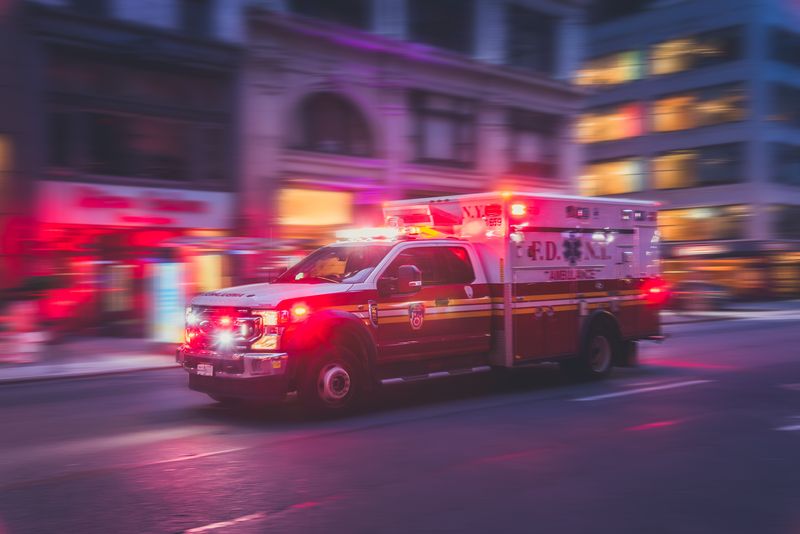 New York ambulance photographed driving fast with red lights on down a road; the background is blurred and the lighting is dark and tinted red