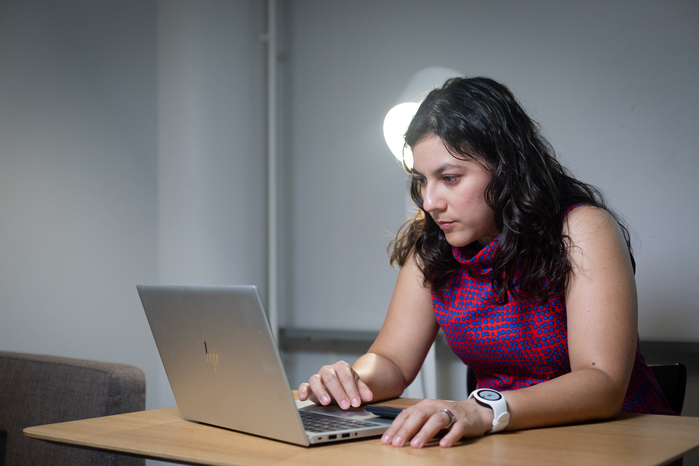 researcher Ana Triana, who has long dark hair and is wearing a red and blue spotted sleeveless dress, sitting at a table using a silver laptop