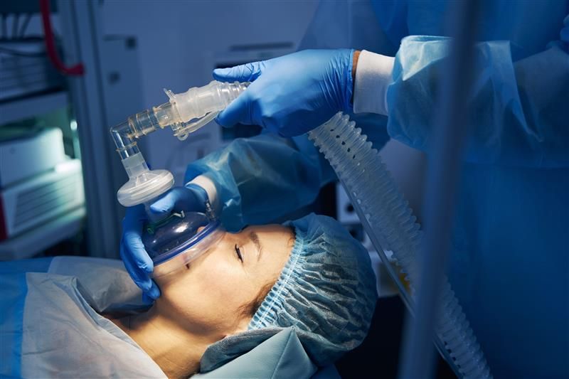 Photo of a woman about to be anesthetised. The shot shows her on profile as the blue rubber mask is being fitted to her mouth and nose. The Anaesthetist's blue gloves hands can be seen holding the mask and the tube connected to it.  