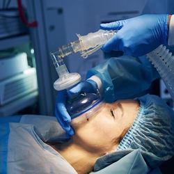 Photo of a woman about to be anesthetised. The shot shows her on profile as the blue rubber mask is being fitted to her mouth and nose. The Anaesthetist's blue gloves hands can be seen holding the mask and the tube connected to it.  