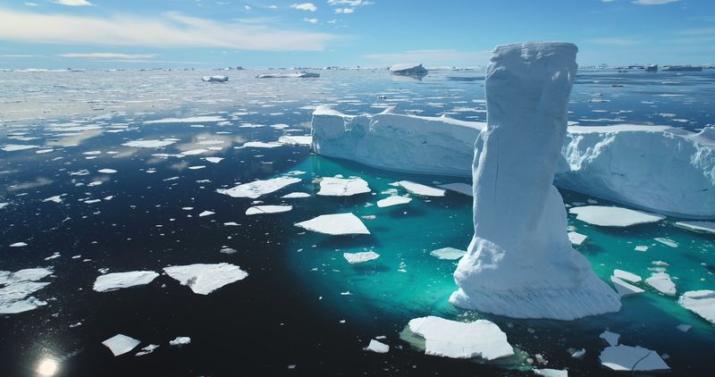 Towering melting iceberg in Arctic icy ocean. Snow covered glacier drift under blue sunny sky. Pure ice floats in turquoise water. Ecology, melting ice, climate change global warming. Aerial view