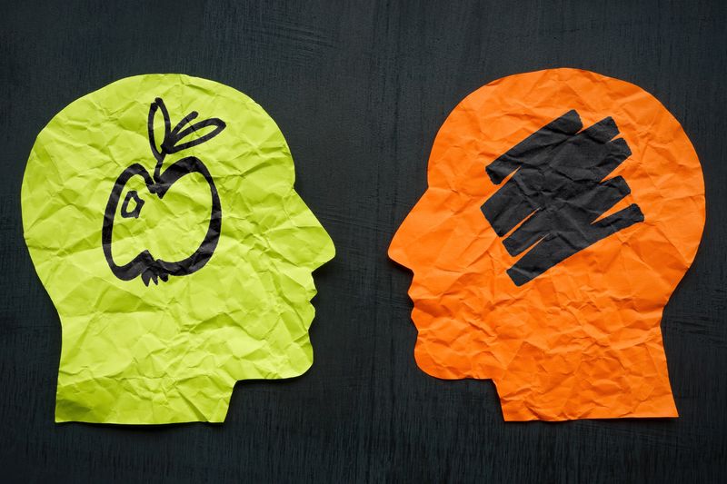 concept art to illustrate aphantasia; two paper silhouettes of a human head face each other on a dark background. The one on the left is lime green and has a drawing of an apple inside in black pen; the one on the left is orange and has a black scribble where the apple should be.