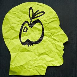 concept art to illustrate aphantasia; two paper silhouettes of a human head face each other on a dark background. The one on the left is lime green and has a drawing of an apple inside in black pen; the one on the left is orange and has a black scribble where the apple should be.