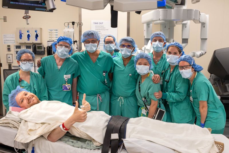 team of surgeons wearing scrubs and masks gathered behind a patient lying in a bed in the OR, giving a thumbs up