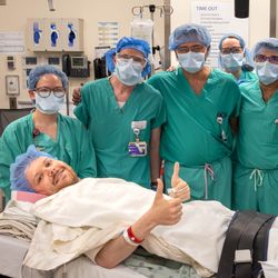 team of surgeons wearing scrubs and masks gathered behind a patient lying in a bed in the OR, giving a thumbs up