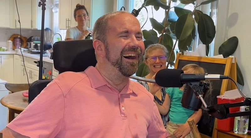 Patient Casey Harrell, a white man with close-shaved hair and a short beard, wearing a pink polo shirt, smiles with his eyes closed  as he tries out the BCI system for the first time. Harrell is wheelchair user and there is a microphone positioned in front of his face