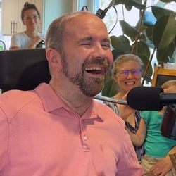 Patient Casey Harrell, a white man with close-shaved hair and a short beard, wearing a pink polo shirt, smiles with his eyes closed  as he tries out the BCI system for the first time. Harrell is wheelchair user and there is a microphone positioned in front of his face