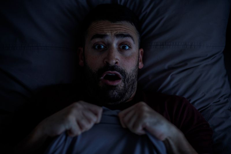 A man in a burgundy T-shirt is scared in bed looking at the camera