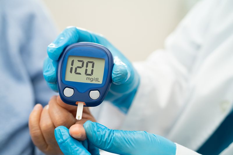 close up of blood glucose monitor showing a reading of 120 mg/dL; a doctor with blue gloves is holding the monitor and taking the blood sample from the patient's finger