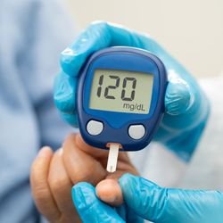 close up of blood glucose monitor showing a reading of 120 mg/dL; a doctor with blue gloves is holding the monitor and taking the blood sample from the patient's finger