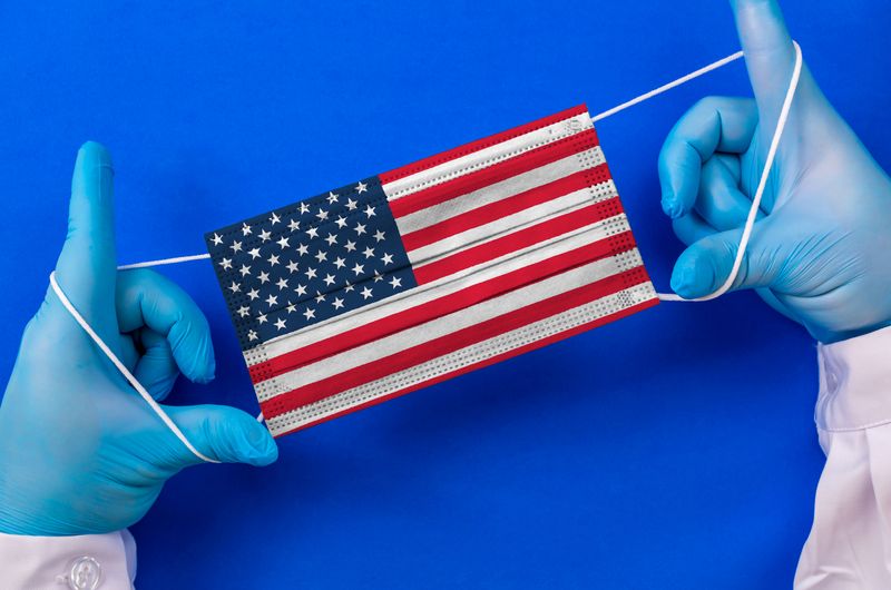 Blue-gloved hands stretching out a surgical mask printed with the USA flag