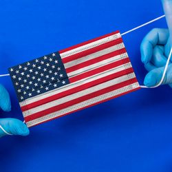 Blue-gloved hands stretching out a surgical mask printed with the USA flag