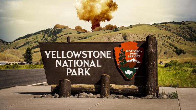 A photo showing the Yellowstone National Park welcome sign with a nuclear mushroom cloud in the background. The photo shows rolling hills in the midground behind the sign which are green and populated with a few trees. The mushroom cloud is peaking up from behind the hills.  