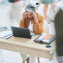 A middle aged woman sits at her desk with her hands pressed to the sides of her head. She is wearing a mustard colored jacket and a white shirt. Her desk has an open laptop in front of her and clipboards and paperwork to either side. There are other people walking around in the image, their bodies are blurred to give the impression that the world is moving quickly while the woman is struggling to focus. 