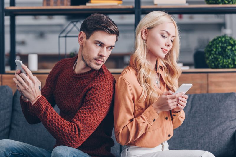 Brown-haired man in red sweater looking suspiciously at the phone of a blonde woman in a brown shirt