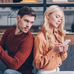 Brown-haired man in red sweater looking suspiciously at the phone of a blonde woman in a brown shirt