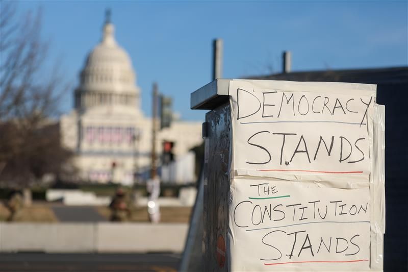 A close up photo of a hand written message on paper attached to a public feature with Capitol Hill in the background. The sign says "Democracy Stands. The Constitution Stands". 