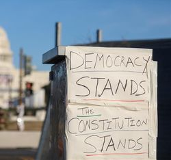 A close up photo of a hand written message on paper attached to a public feature with Capitol Hill in the background. The sign says "Democracy Stands. The Constitution Stands". 