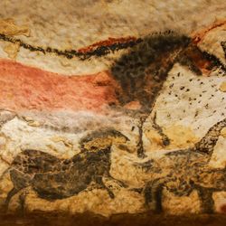 A photo showing the cave art at the Lascaux Cave in France. The image shows a horse painted in red medium being imposed on the black outline of a large bull-like animal. Below it are smaller black representations of other horse-like animals. 