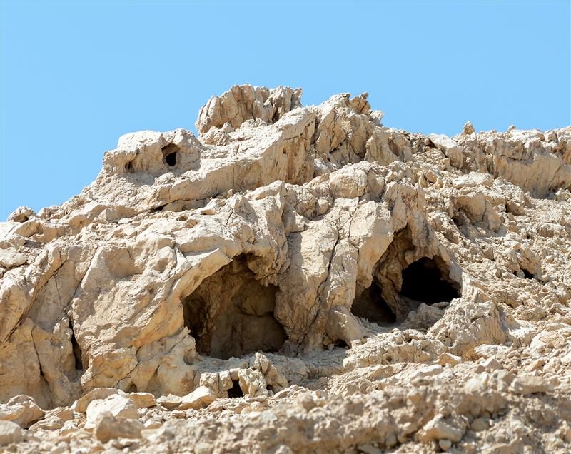 A photo of a series of caves in a rocky face of a cliff. The image shows two of the caves surrounding by craggy pale rocks. The scene is completely absent of any vegetation.  