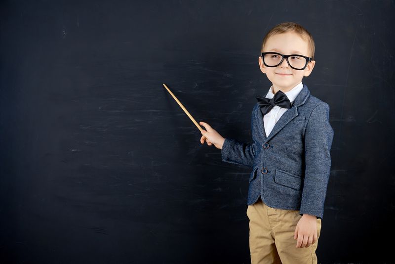 Child dressed in a tweed jacket, glasses, and a bow tie, pointing stick at blackboard