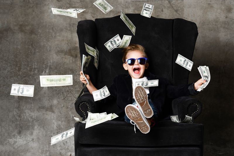 Child in sunglasses laughing and sitting in a black armchair, throwing cash in the air