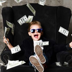 Child in sunglasses laughing and sitting in a black armchair, throwing cash in the air