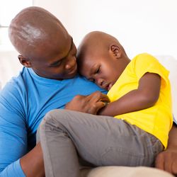 Child sitting on father's lap, being comforted. Both are sat on a white couch.