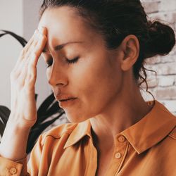 woman with eyes closed holding hand to her forehead to indicate fatigue; she is wearing an orange blouse and there's a brick wall and a plant in the background