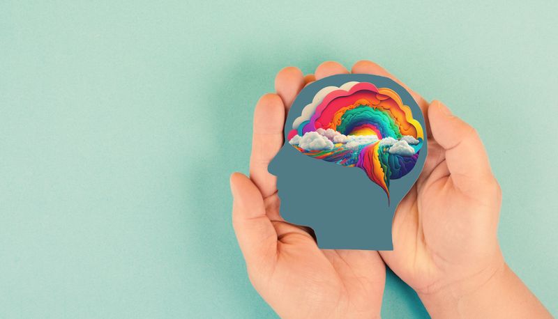 hands holding silhouette of human head with rainbow colored brain against light aqua background