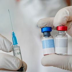 concept image for a combined vaccine, close up of doctor holding two vaccine vials and a syringe in white-gloved hands