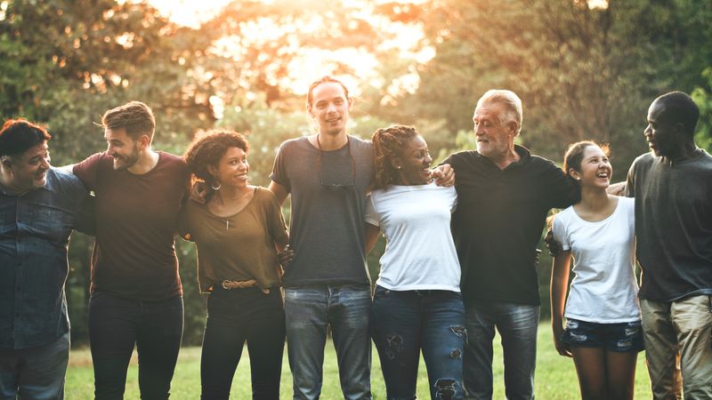 A photo of people lined up arm in arm to form a chain. The line of people is made up of individuals of different sex, age and race and the are all smiling and enjoying themselves. They are in what appears to be a garden with a green hedge background and the setting sun shining through the leaves.  