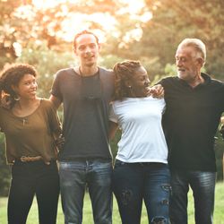 A photo of people lined up arm in arm to form a chain. The line of people is made up of individuals of different sex, age and race and the are all smiling and enjoying themselves. They are in what appears to be a garden with a green hedge background and the setting sun shining through the leaves.  