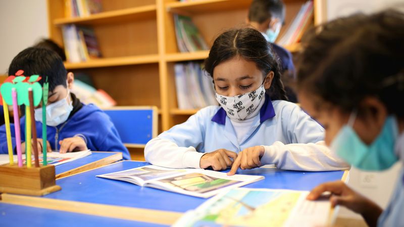 three children wearing face masks sitting at a desk at school reading