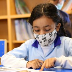 three children wearing face masks sitting at a desk at school reading