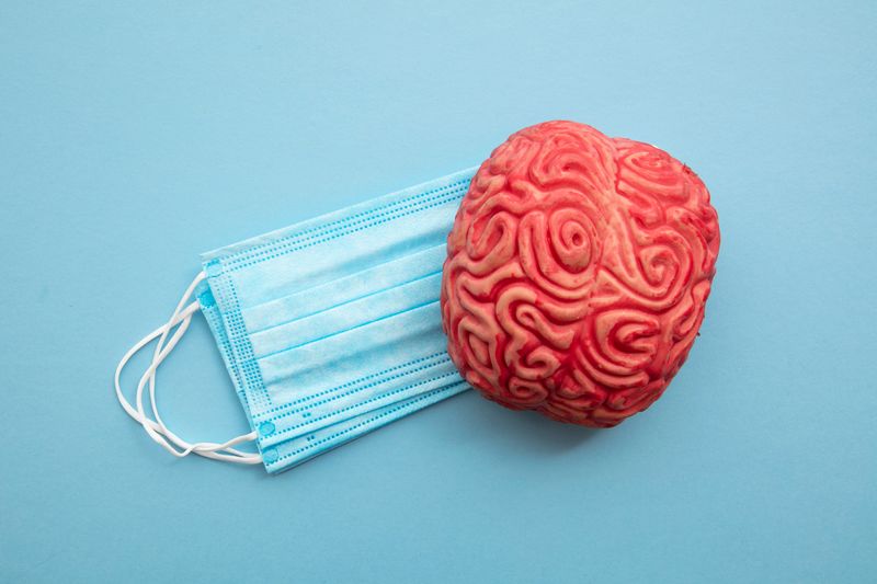 flat lay on pale blue surface; there is a small stack of blue surgical masks with a red 3D model of the human brain slightly overlaying them to the right hand side