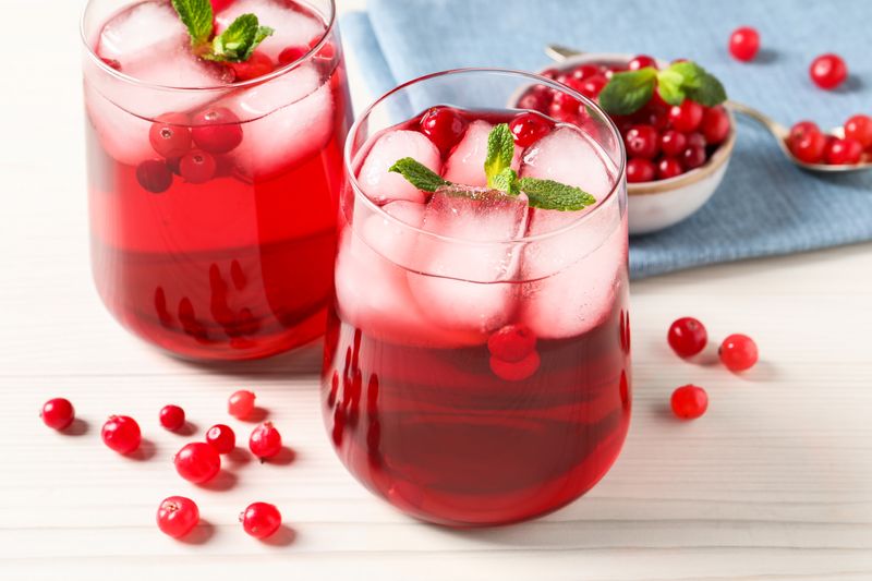 two small glasses of cranberry juice with ice cubes in them, placed on a white wooden table with cranberries also scattered on it