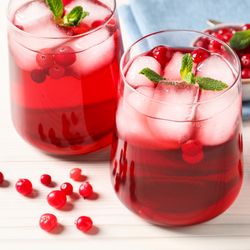 two small glasses of cranberry juice with ice cubes in them, placed on a white wooden table with cranberries also scattered on it