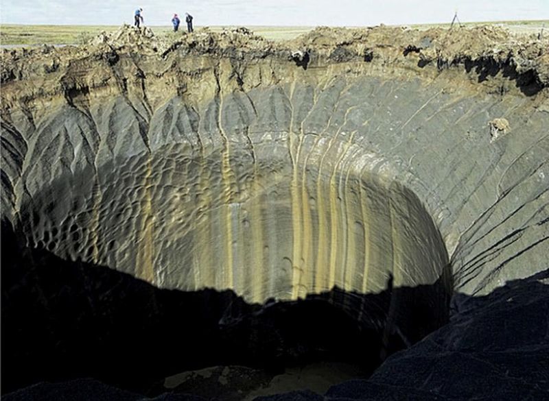 A photo shows the enormous crater caused by the explosions. The hole has a lip of grey clay that then drops into a deep shaft that is tends of feet across. In the background of the image, three people are standing looking into the hole but they are tiny compared to its size.