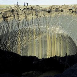 A photo shows the enormous crater caused by the explosions. The hole has a lip of grey clay that then drops into a deep shaft that is tends of feet across. In the background of the image, three people are standing looking into the hole but they are tiny compared to its size.