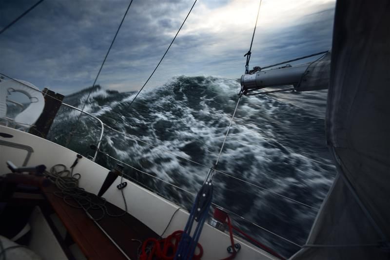 A photo taken from a deck of a yacht one a turbulent sea. The vessel is tipped to the right and the waves are rising above its eyeline. The is grey and cloudy, but there is light in the top right hand corner.  