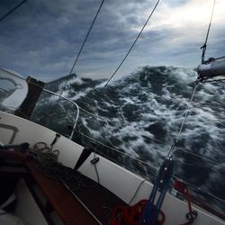 A photo taken from a deck of a yacht one a turbulent sea. The vessel is tipped to the right and the waves are rising above its eyeline. The is grey and cloudy, but there is light in the top right hand corner.  